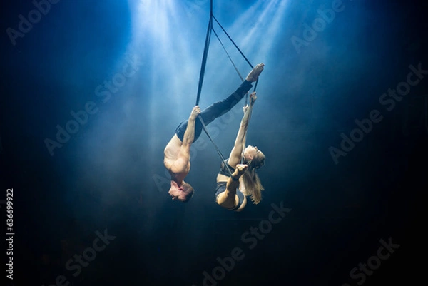 Fototapeta An aerial straps duo performs dangerous tricks mid-air, illuminated by strong blue and white lights against a dark background, clad in minimal athletic attire, showcasing a blend of power and grace