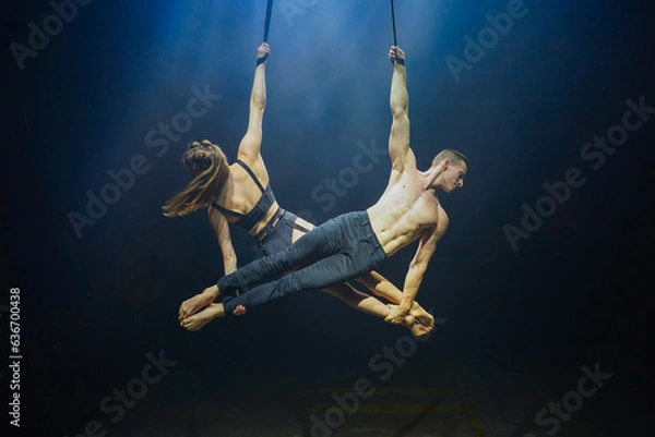 Fototapeta Aerial straps duo performance: a man and woman execute graceful acrobatic feats in mid-air against a black background, dressed in black and illuminated by a white-blue glow