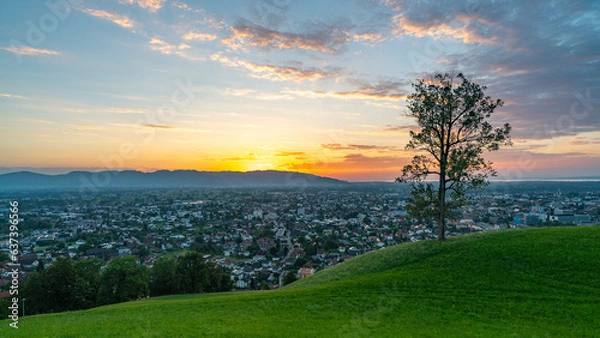 Fototapeta Panorama Sonnenuntergang über den Schweizer Bergen im Rheintal, einzelner Baum mit Blick auf Dornbirn, Vorarlberg, Austria. leuchtendes Abendrot, tolle Stimmung mit roten Wolken über den grünen Wiesen