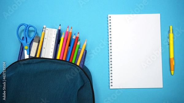 Fototapeta School supplies looks out from school bag near open student notebook with yellow pen on blue background. Back to school, education, learning concept. Top view