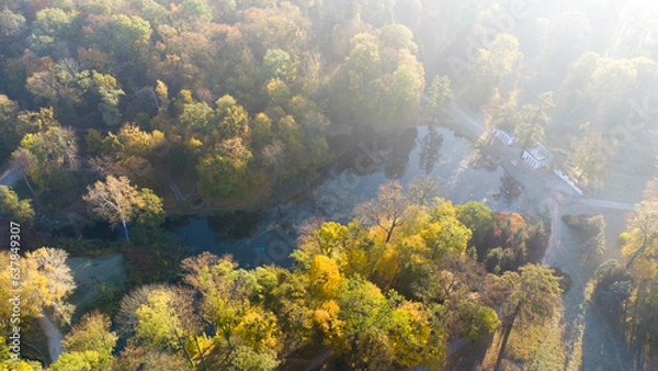 Fototapeta Aerial flying over trees with yellow leaves, lake, columns and walking people on autumn sunny morning in park. Bright sunlight, shining sunbeams rays and sun overexposure. Beautiful natural background
