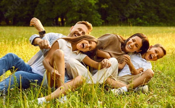 Fototapeta Happy young family of four smiling and having fun while sitting on green grass in park during summer rest. Laughing mother and father hugging their kids boy and girl lying on the field.