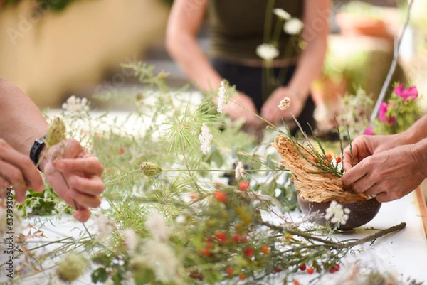 Fototapeta Detail of hands making flower arrangement, outdoor ikebana workshop, different flower arrangements in progress.
