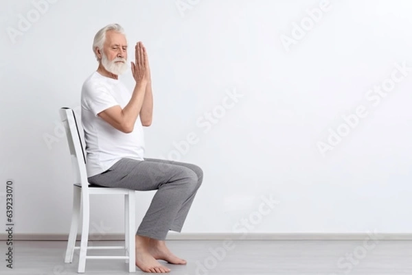 Obraz An elderly man practices yoga asana or sports exercises for legs and arms on a chair. The concept of sports in old age, exercise therapy, rehabilitation. Banner. Copy space