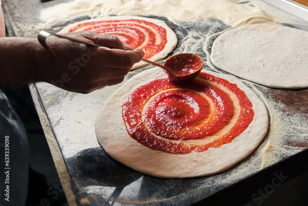 Obraz A man spreads Italian tomato paste over the dough, part of the pizza preparation process at the family café