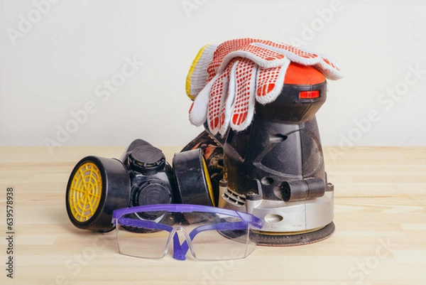 Fototapeta Grinding machine, gloves, goggles and a protective mask on a wooden table against a white wall.