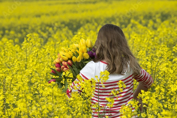 Fototapeta Child walking in a field of rapeseed holding Spring flowers