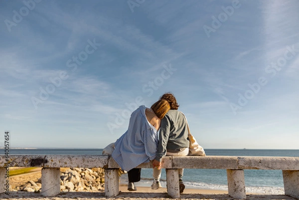Fototapeta Rear view of couple hugs while sitting on bench and enjoys beautiful ocean view. High quality photo