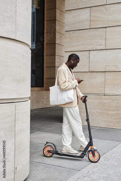 Fototapeta Long shot of African American businessman with scooter, smartphone and white textile handbag standing by modern architecture and texting