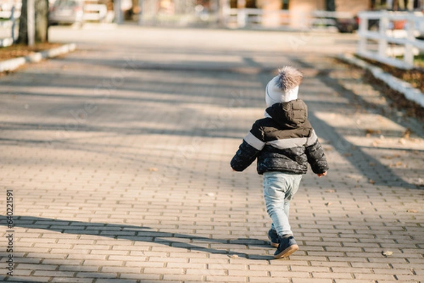 Fototapeta Child walking in park at sunset. Family spending time on vacation. Back view. Kid runs on street in nature. Concept of autumn holiday. Portrait of baby, toddler. Space for text. Place for advertising.