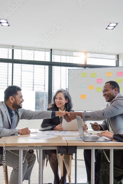 Fototapeta African professional businessman and Indian male successful businessman in formal suit sitting shaking hands when business agreement deal done while female colleagues clapping hands celebrating.
