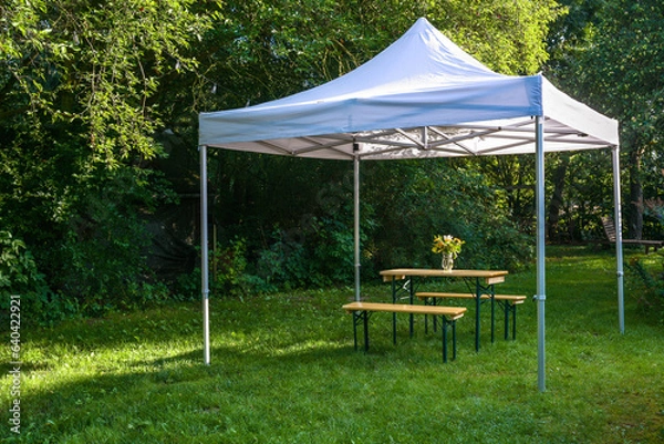 Fototapeta White gazebo or pavilion with a wooden table, benches and a flower bouquet on the meadow in a natural garden, idyllic summer place gives protection from sun and rain, copy space