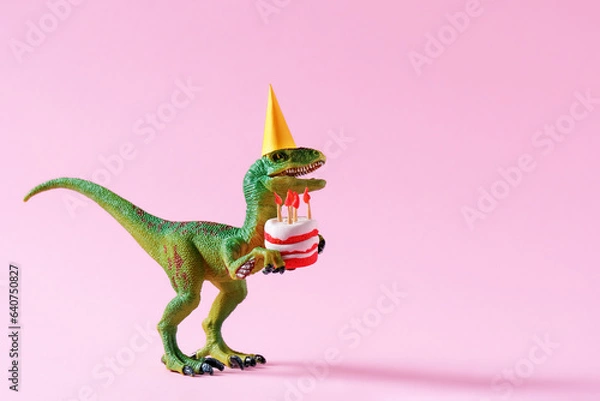 Fototapeta Cute happy green dinosaur in birthday hat holding cake with flaming candles on pastel pink background. Copy space. Minimal art birthday card idea.