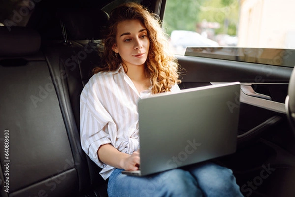Fototapeta Stylish woman with glasses in the car, sitting on the back seat of the car and working on a laptop. Business concept, blogging, freelancing.