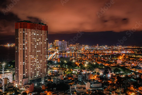 Fototapeta Nha Trang from the drone in the evening. In the background there is a skyscraper and many illuminated buildings. Orange clouds in the sky. Vietnam