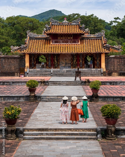 Fototapeta Hue, Vietnam, Hien Duc Gate At Mausoleum Of Minh Mang. . Against the background of the mausoleum, three girls in traditional Vietnamese costumes are standing with their backs.
