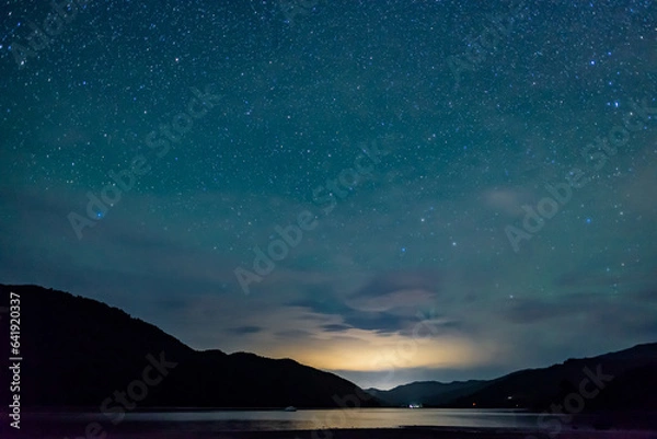 Obraz starry sky above the shape of mountains with the light of a city in a fjord in the Marlborough Sounds, New Zealand