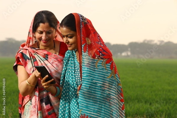Obraz Happy rural Indian women standing in a mustard agricultural field and surfing through the mobile phone in their hand.