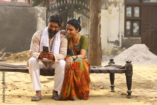 Obraz Happy Indian Rural family in village. couple sitting together outside their home front yard using mobile phone. Wife beautiful saree traditional wear