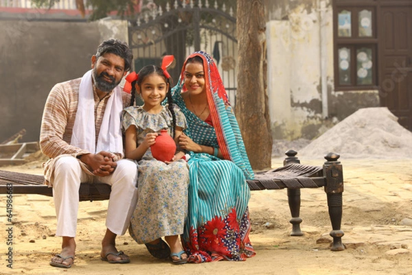 Obraz Happy rural Indian family sitting together, daughter is holding a piggy bank in her hand, putting a coin in it, showing the concept of saving money