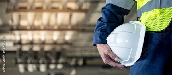 Obraz Safety workwear concept. Male hand holding white safety helmet or hard hat. Construction worker man in protective suit and reflective green vest standing with building piping system in the background