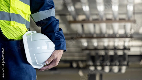 Obraz Safety workwear concept. Male hand holding white safety helmet or hard hat. Construction worker man in protective suit and reflective green vest standing with building piping system in the background
