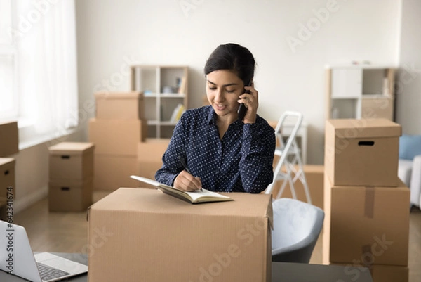 Fototapeta Pretty young Indian storage manager girl sending purchase from Internet store to customer, talking on mobile phone at packed cardboard box, writing notes, processing order