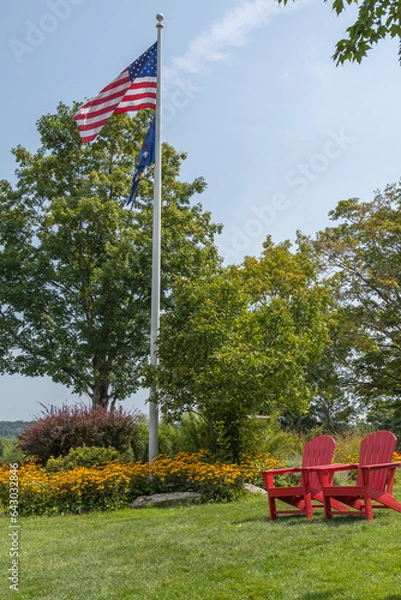 Obraz Flag pole with American flag flying and 2 red deck chairs in outdoor setting. 