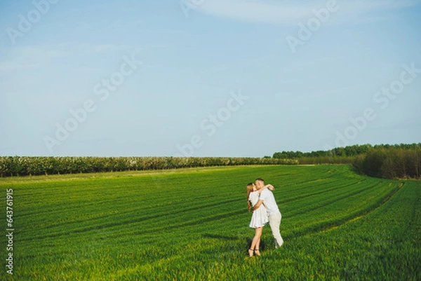 Fototapeta Pregnant couple hugging in a green spring field. Expectant parents in the park of white flowering trees. A romantic couple expecting a baby. A walk through a green field