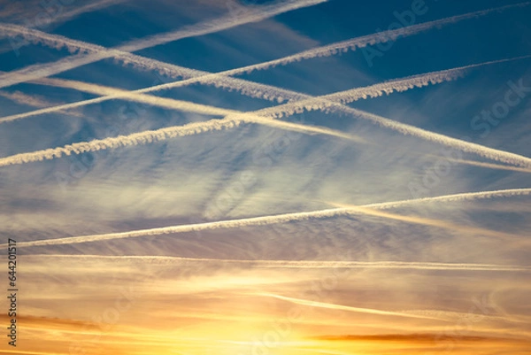 Fototapeta Around 10 airplane vapor trails in the sky in a diagonal pattern against a dark blue sky at sunset with yellow and orange clouds on the horizon.