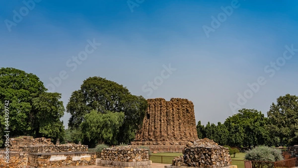 Fototapeta The ancient unfinished brick minaret Ala-i-Minar in the temple complex Qutb-minar. The first tier of the tower with weathered walls against the blue sky. Green vegetation around. India. Delhi.