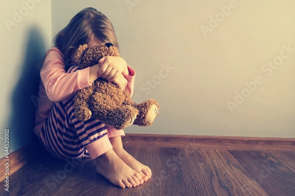 Fototapeta The child is sitting sad scared on the floor in the corner and hugs a toy. Little girl crying with her head down. Concept: No Domestic Violence and Child Abuse. Mistakes of parents in raising children