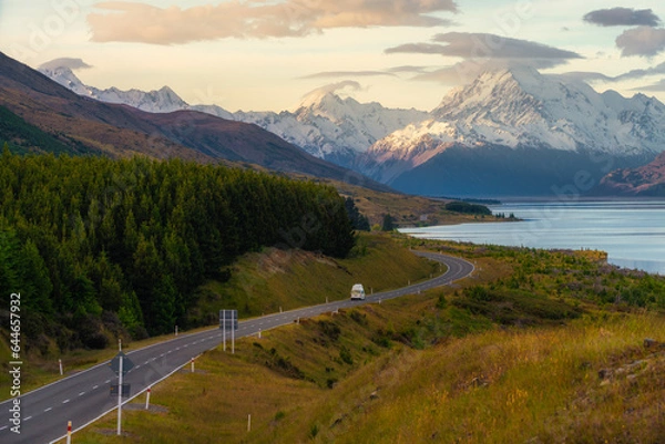 Fototapeta A car driving along a winding road beside a lake with snow capped mountains in the background.