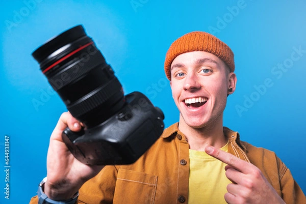 Fototapeta Happy smiling young  photographer taking pictures checking his camera settings in a studio shooting as content creator in blue background 
