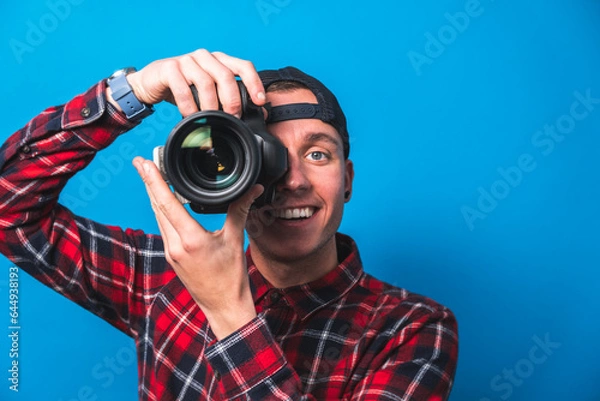 Fototapeta Happy photographer taking pictures checking his camera settings in a studio shooting as content creator in blue background 