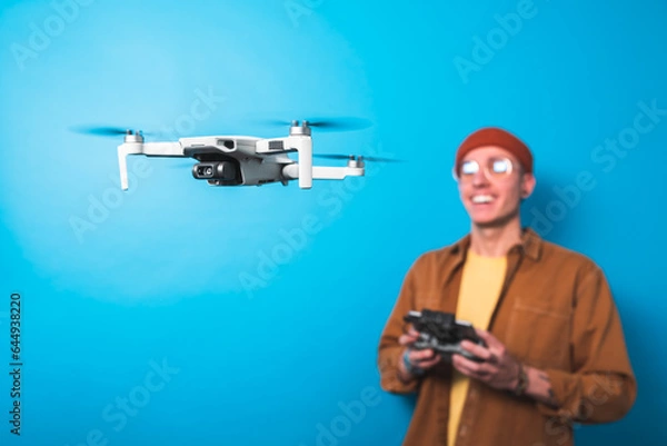 Fototapeta Smiling young man piloting a drone in flight with a remote control on an isolated blue background. Drone pilot and aerial filming concepts. Focus on the drone