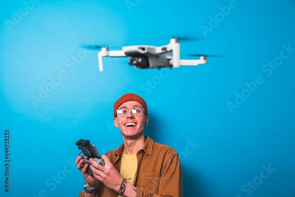 Fototapeta Smiling young man learning piloting a drone in flight with a remote control on an isolated blue background. Drone pilot and aerial filming concepts. Focus on the drone