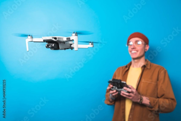 Fototapeta Smiling young man training piloting a drone in flight with a remote control on an isolated blue background. Drone pilot and aerial filming concepts. 