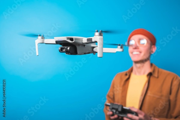 Fototapeta Smiling young man piloting a drone in flight with a remote control on an isolated blue background. Drone pilot and aerial filming concepts. Focus on the drone