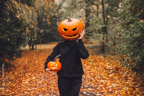 Fototapeta Halloween. Pumpkinhead. A man with a pumpkin on his head stands in a park on an alley strewn with yellow autumn leaves.