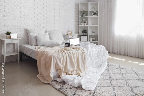 Fototapeta Messy bed with white blanket, coffee on tray and modern laptop in light bedroom