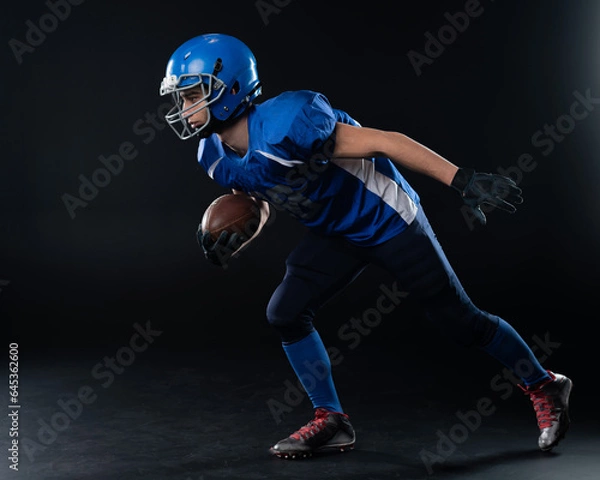 Fototapeta Full length portrait of a man in a blue american football uniform against a black background. Sportsman in a helmet with a ball. 