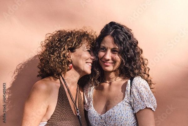 Fototapeta Mother and daughter posing smiling and affectionate in front of a brown wall.