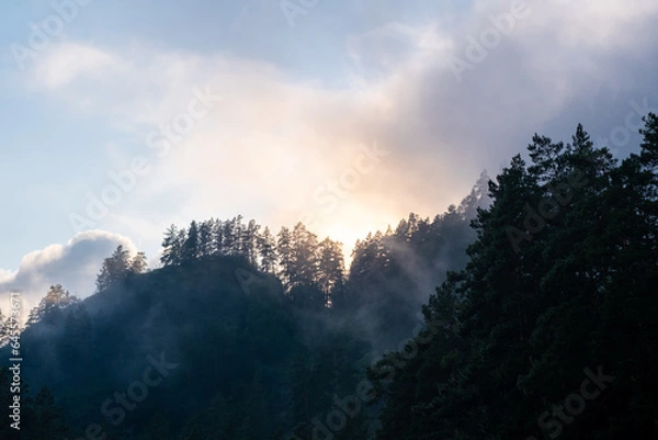 Obraz Mountains and peaks in fog at sunrise or sunset. Sun from behind the mountain. Amazing view of mountains and forest landscape with cloudy skies Altai mountains. Mountains and peaks in fog at sunrise