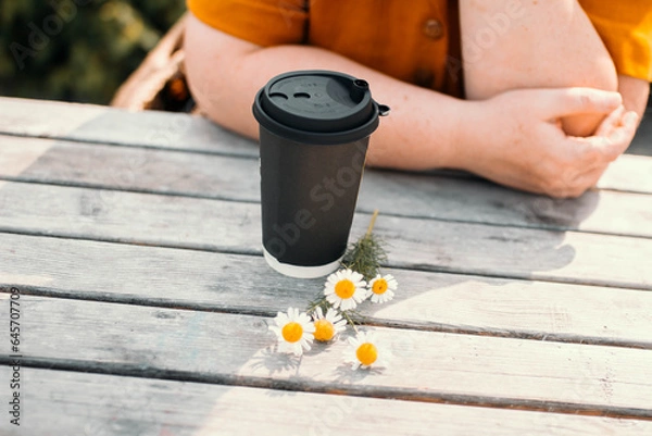 Obraz Black paper cup for coffee or tea standing on a wooden table with a sprig of daisies and woman's hands in the background