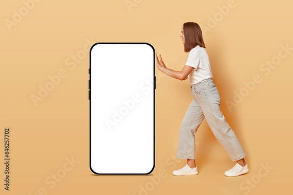 Fototapeta App service. Social media influencer. Online shopping experience. Happy cheerful brown haired adult woman pulling big phone with white empty display isolated over beige background