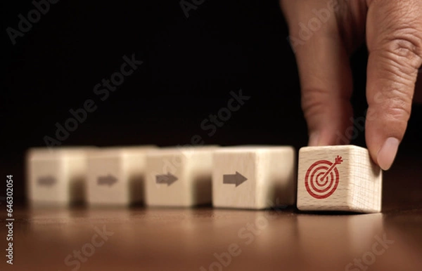 Fototapeta Arrow on wooden block pointing at target icon on wooden block with businessman's hand placing red dartboard symbol. Business Agenda and Mission Concepts Cube dartboard. Future vision strategy, goal, s