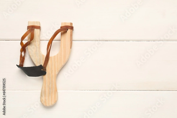 Fototapeta Slingshot with leather pouch on white wooden background, top view. Space for text