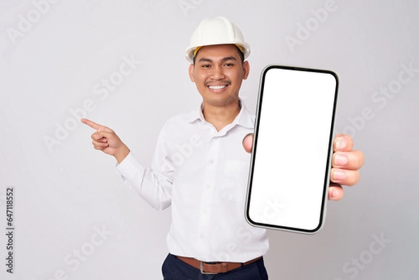 Fototapeta Smiling young Asian engineer employee man in a formal shirt helmet holding smartphone with blank screen and pointing finger aside isolated on white background. People work on architecture project