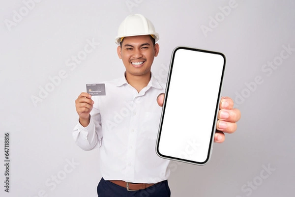 Fototapeta Smiling young Asian architect engineer employee man in a formal shirt helmet holding smartphone with blank screen and credit card isolated on white background. People work on architecture project